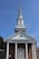 A traditional church building with a tall steeple against a clear blue sky, featuring a brick facade and white columns. The words 'CHURCH OF CHRIST' are prominently displayed above the entrance.
