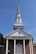 A traditional church building with a tall steeple against a clear blue sky, featuring a brick facade and white columns. The words 'CHURCH OF CHRIST' are prominently displayed above the entrance.