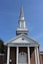 A traditional church building with a tall steeple against a clear blue sky, featuring a brick facade and white columns. The words 'CHURCH OF CHRIST' are prominently displayed above the entrance.