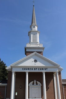 A traditional church building with a tall steeple against a clear blue sky, featuring a brick facade and white columns. The words 'CHURCH OF CHRIST' are prominently displayed above the entrance.