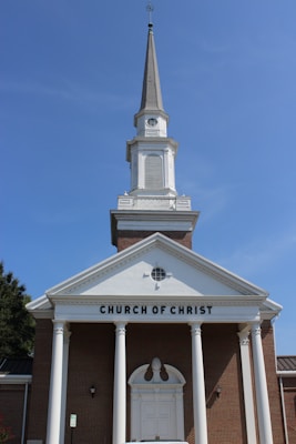 A traditional church building with a tall steeple against a clear blue sky, featuring a brick facade and white columns. The words 'CHURCH OF CHRIST' are prominently displayed above the entrance.