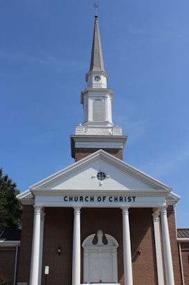 A traditional church building with a tall steeple against a clear blue sky, featuring a brick facade and white columns. The words 'CHURCH OF CHRIST' are prominently displayed above the entrance.