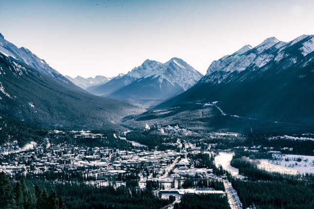 A scenic view captures a mountainous landscape with snow-capped peaks and a forested valley. A small town is nestled in the valley, with buildings scattered amongst the trees, and a clear road cutting through the area. The sky is bright with a hint of a hazy sun.