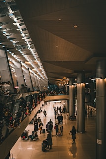 A spacious, modern interior with a high ceiling and polished floors. People are walking and pulling luggage, indicating a travel or transit area like an airport. Christmas decorations with lights and garlands line a glass railing, creating a festive atmosphere.