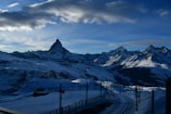 A mountain landscape features snow-covered peaks with a prominent jagged summit under a bright blue sky with scattered clouds. Below, a railway track winds through the snowy terrain.