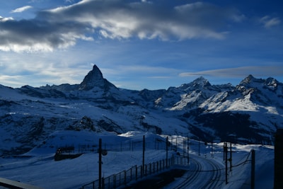 A mountain landscape features snow-covered peaks with a prominent jagged summit under a bright blue sky with scattered clouds. Below, a railway track winds through the snowy terrain.