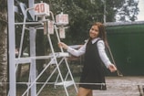 A young woman in a black dress and white blouse is smiling while standing next to a scoreboard outdoors. The scoreboard displays the numbers 40 and 0. There is greenery and a green building in the background.