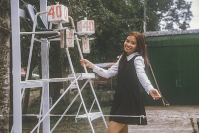 A young woman in a black dress and white blouse is smiling while standing next to a scoreboard outdoors. The scoreboard displays the numbers 40 and 0. There is greenery and a green building in the background.