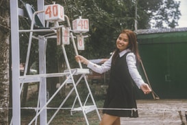 A young woman in a black dress and white blouse is smiling while standing next to a scoreboard outdoors. The scoreboard displays the numbers 40 and 0. There is greenery and a green building in the background.