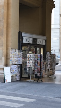 A newsstand situated under a large stone arch, featuring racks filled with various magazines, postcards, and newspapers. The word 'VOGUE' is prominently displayed above the stand. The surrounding architecture consists of large beige stone columns.