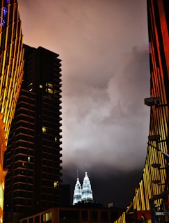 The image captures a nighttime cityscape with the Petronas Twin Towers illuminated in the background against a cloudy sky. Tall buildings frame the scene on both sides, with lights creating a dramatic contrast against the dark atmosphere.
