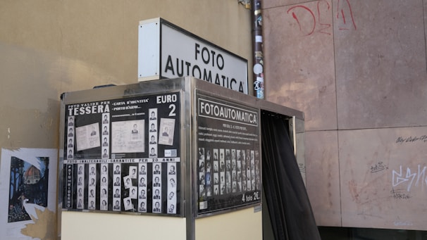A vintage photo booth called Fotoautomatica is positioned against a graffiti-marked wall. The booth is surrounded by posters displaying sample passport photos and information in Italian, indicating a cost of two Euros for obtaining photos. The booth exterior has a metallic finish and a black curtain that conceals the interior.
