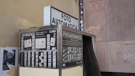 A vintage photo booth called Fotoautomatica is positioned against a graffiti-marked wall. The booth is surrounded by posters displaying sample passport photos and information in Italian, indicating a cost of two Euros for obtaining photos. The booth exterior has a metallic finish and a black curtain that conceals the interior.