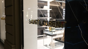 A high-end fashion store display with elegant black and white marble accents. The storefront features the Saint Laurent logo in gold lettering on a reflective glass surface. Inside the store, luxury handbags are displayed on sleek, illuminated shelves and glass tables.