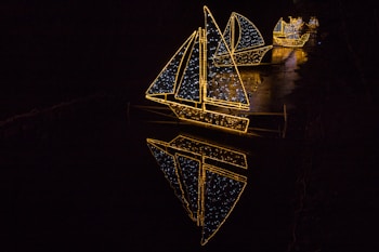 A series of sailboats outlined with strings of bright yellow and white lights display their reflections in the dark water below. The scene is set against a pitch-black background, emphasizing the vivid illumination of the sails and hulls.