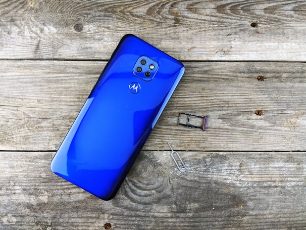 A bright blue smartphone with four cameras on the back is placed on a textured wooden surface. Nearby, there is a SIM card tray and a SIM card removal tool.