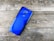 A bright blue smartphone with four cameras on the back is placed on a textured wooden surface. Nearby, there is a SIM card tray and a SIM card removal tool.