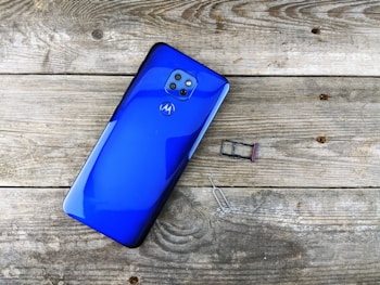 A bright blue smartphone with four cameras on the back is placed on a textured wooden surface. Nearby, there is a SIM card tray and a SIM card removal tool.