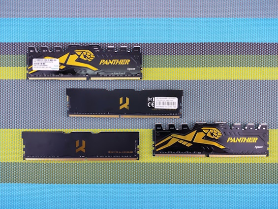 Four black computer RAM modules with gold and yellow accents are arranged on a textured, multicolored surface. Two of the modules have the word 'PANTHER' and an illustrated panther logo on them, while the other two have a stylized logo in the center. All modules show technical details and barcodes on stickers.