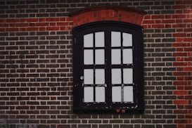 Black-framed window with multiple panes set in a brick wall. The brickwork around the window has a distinct pattern, with red bricks forming an arch above the window. The window appears weathered, with signs of chipping paint.