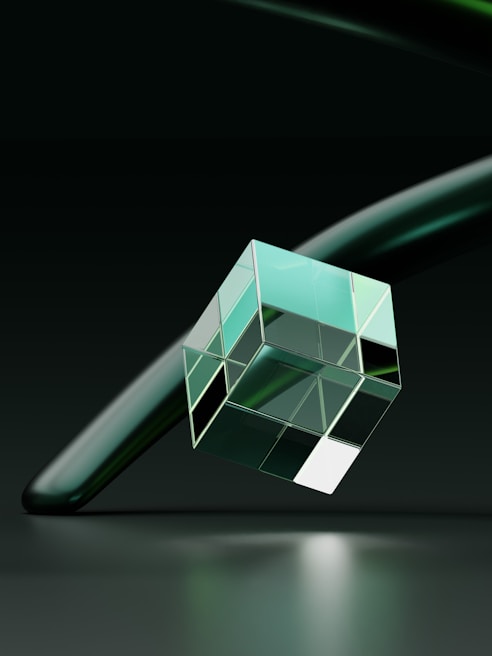 A transparent geometric cube with reflective surfaces is suspended in the air. The background features a smooth, dark, and slightly blurred surface, with another elongated, curved object crossing behind the cube. The lighting casts soft reflections and highlights on the surfaces of the cube.