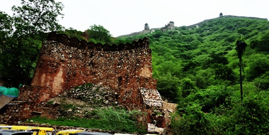 An ancient stone fort with tall, rugged walls stands against a backdrop of lush green hills. The fort exhibits a reddish-brown hue with visible stonework and irregular shapes. A dense growth of trees and greenery covers the hillside, contributing to the natural setting. At the base of the image, several auto rickshaws are visible, indicating modern activity near the historic structure.