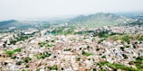 An aerial view of a densely populated town surrounded by lush greenery and hilly terrain in the background. The town consists of numerous buildings closely packed together, with a variety of architectural styles. Green foliage is interspersed among the buildings, and the horizon features rolling hills covered with vegetation under a cloudy sky.