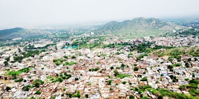 An aerial view of a densely populated town surrounded by lush greenery and hilly terrain in the background. The town consists of numerous buildings closely packed together, with a variety of architectural styles. Green foliage is interspersed among the buildings, and the horizon features rolling hills covered with vegetation under a cloudy sky.