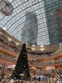 A spacious shopping mall interior decorated for the holiday season, featuring a large Christmas tree adorned with ornaments and lights. The mall has multiple levels with various stores and a glass ceiling that reveals the outlines of tall buildings outside. There are escalators and festive decorations throughout.