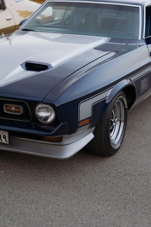 A vintage muscle car with a dark blue body and white stripes, featuring a distinctive hood scoop and chrome wheels. The car is parked on a light-colored pavement, with part of another vehicle visible in the background.