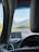 View from inside a car looking through the windshield at a scenic highway with mountains in the background. A GPS device is mounted on the dashboard showing directions. The sky is clear with a few clouds and there are trees lining the road on both sides.
