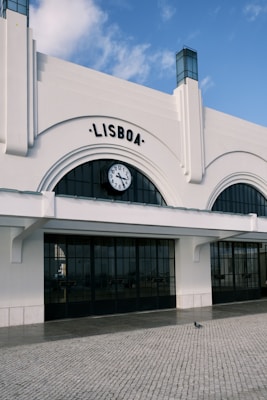A building with an arched facade and the word 'Lisboa' displayed above a large clock. The structure features white walls and black-framed windows. The ground is cobblestone, and a single pigeon is visible in the foreground. The sky is partly cloudy, hinting at a clear day.