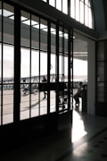 The image features a large industrial-style glass door with a grid of panes, casting intricate shadows on the polished floor. Sunlight floods through the glass, illuminating the interior and highlighting the textures of the stone floor. Outside, a few people are walking on a paved area, with structural elements of a modern building visible.