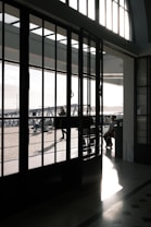 The image features a large industrial-style glass door with a grid of panes, casting intricate shadows on the polished floor. Sunlight floods through the glass, illuminating the interior and highlighting the textures of the stone floor. Outside, a few people are walking on a paved area, with structural elements of a modern building visible.