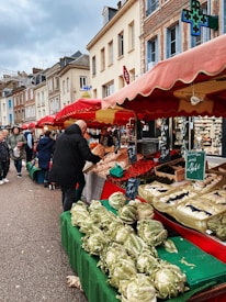 A bustling outdoor market scene with various stalls under red canopies. Displays of fresh vegetables like cabbages and radishes are arranged on tables covered with green cloths. People are walking and interacting with vendors amidst a backdrop of charming European buildings.