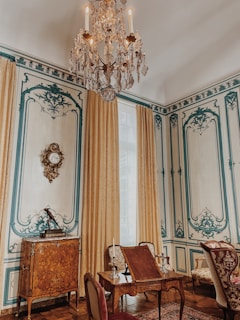 An elegantly decorated room featuring intricate wall designs with blue accents and beige tones. A large, ornate chandelier hangs from the ceiling, while richly embroidered curtains frame a tall window. A wooden cabinet with elaborate inlays and a decorative clock is against the wall. In the foreground, there is a set of antique wooden furniture with upholstered chairs and a small table adorned with candelabras.