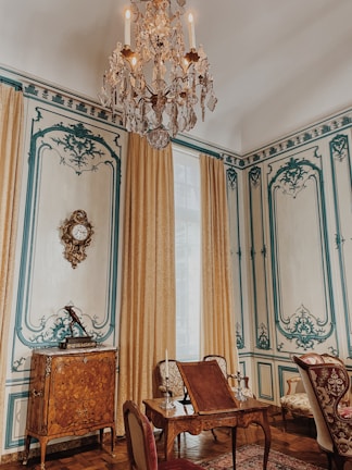 An elegantly decorated room featuring intricate wall designs with blue accents and beige tones. A large, ornate chandelier hangs from the ceiling, while richly embroidered curtains frame a tall window. A wooden cabinet with elaborate inlays and a decorative clock is against the wall. In the foreground, there is a set of antique wooden furniture with upholstered chairs and a small table adorned with candelabras.
