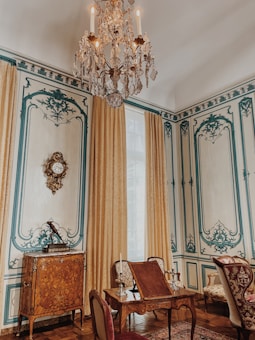 An elegantly decorated room featuring intricate wall designs with blue accents and beige tones. A large, ornate chandelier hangs from the ceiling, while richly embroidered curtains frame a tall window. A wooden cabinet with elaborate inlays and a decorative clock is against the wall. In the foreground, there is a set of antique wooden furniture with upholstered chairs and a small table adorned with candelabras.