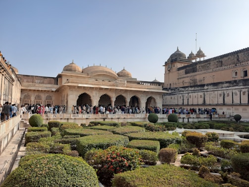 A historic palace with ornate architectural features, including domes and arches, in a clear blue sky setting. The foreground showcases manicured gardens with neatly trimmed hedges and pathways. A large group of people are gathered near the palace, suggesting it is a tourist attraction.