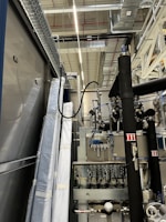 An industrial setting with machinery, visible ductwork, and metal equipment. Pipes and cables are interconnected, and some components are wrapped in protective material. The ceiling features a grid of lights and HVAC ducts.