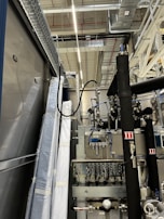 An industrial setting with machinery, visible ductwork, and metal equipment. Pipes and cables are interconnected, and some components are wrapped in protective material. The ceiling features a grid of lights and HVAC ducts.
