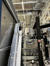 An industrial setting with machinery, visible ductwork, and metal equipment. Pipes and cables are interconnected, and some components are wrapped in protective material. The ceiling features a grid of lights and HVAC ducts.