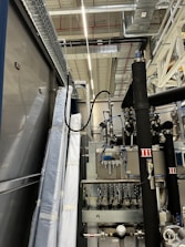 An industrial setting with machinery, visible ductwork, and metal equipment. Pipes and cables are interconnected, and some components are wrapped in protective material. The ceiling features a grid of lights and HVAC ducts.