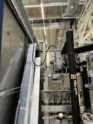 An industrial setting with machinery, visible ductwork, and metal equipment. Pipes and cables are interconnected, and some components are wrapped in protective material. The ceiling features a grid of lights and HVAC ducts.