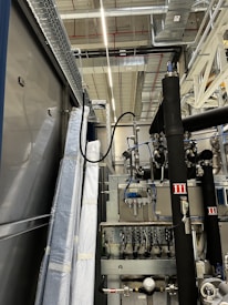 An industrial setting with machinery, visible ductwork, and metal equipment. Pipes and cables are interconnected, and some components are wrapped in protective material. The ceiling features a grid of lights and HVAC ducts.