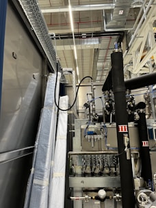 An industrial setting with machinery, visible ductwork, and metal equipment. Pipes and cables are interconnected, and some components are wrapped in protective material. The ceiling features a grid of lights and HVAC ducts.
