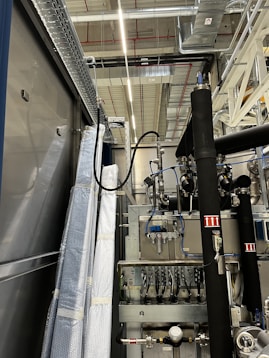 An industrial setting with machinery, visible ductwork, and metal equipment. Pipes and cables are interconnected, and some components are wrapped in protective material. The ceiling features a grid of lights and HVAC ducts.