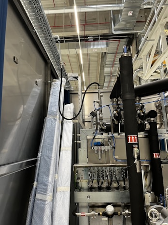 An industrial setting with machinery, visible ductwork, and metal equipment. Pipes and cables are interconnected, and some components are wrapped in protective material. The ceiling features a grid of lights and HVAC ducts.