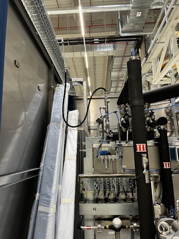 An industrial setting with machinery, visible ductwork, and metal equipment. Pipes and cables are interconnected, and some components are wrapped in protective material. The ceiling features a grid of lights and HVAC ducts.