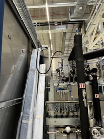 An industrial setting with machinery, visible ductwork, and metal equipment. Pipes and cables are interconnected, and some components are wrapped in protective material. The ceiling features a grid of lights and HVAC ducts.